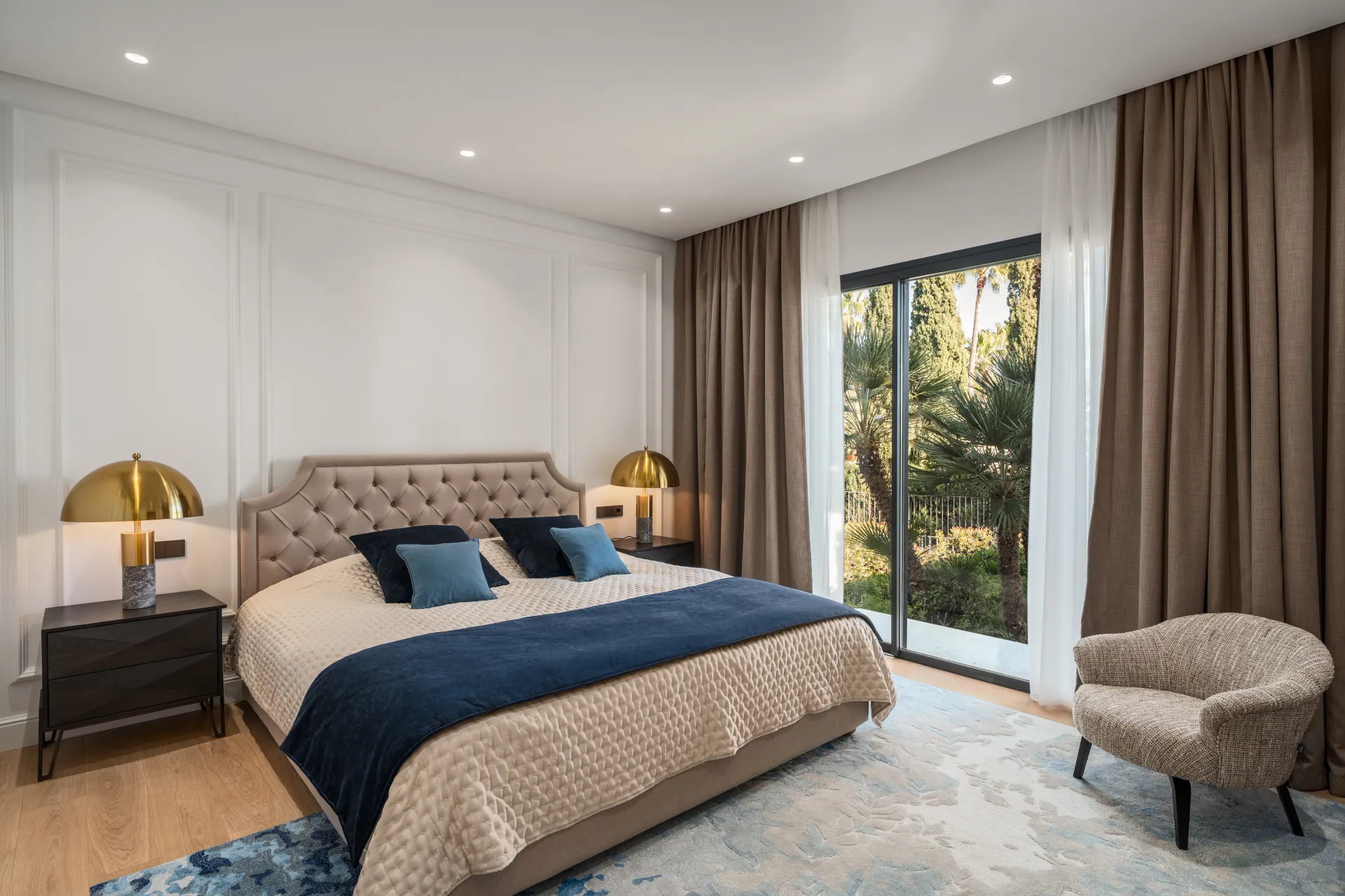 Contrasting calm. Tufted headboard, brass lamp, barrel chair and a view of palm trees. The deep blue accents create a sophisticated dialogue with the surrounding nature.